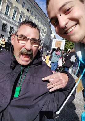 Ein Bewohner und ein Mitarbeiter von BALANCE machen ein fröhliches Selfie auf einer Demonstration für Inklusion in Wien. Im Vordergrund lacht ein Mann mit Brille und Schnurrbart ausgelassen in die Kamera, trägt eine grüne Trillerpfeife um den Hals und zeigt mit der Hand ein Rock-Geste-Symbol. Rechts im Bild lächelt ein jüngerer Mitarbeiter mit Piercings herzlich. Im Hintergrund sind weitere Teilnehmer der Demonstration, Transparente und historische Gebäude unter blauem Himmel zu sehen.