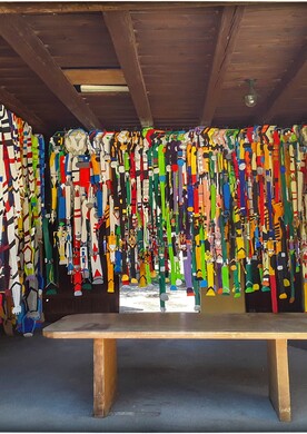 Eine raumgreifende Kunstinstallation von Barbara Plak in einem überdachten Außenbereich des Ateliers Maria Ponsee. Hunderte schmale, vertikale Papiergestalten (Androiden) hängen dicht an dicht von der Decke herab und füllen die drei Wände des Raumes fast vollständig aus. Jeder Streifen ist individuell mit geometrischen Mustern und leuchtenden Farben wie Gelb, Blau, Rot und Grün gestaltet. Die Anordnung erinnert an einen Vorhang aus bunten Bändern, der einen hölzernen Tisch im Zentrum des Raumes umschließt.