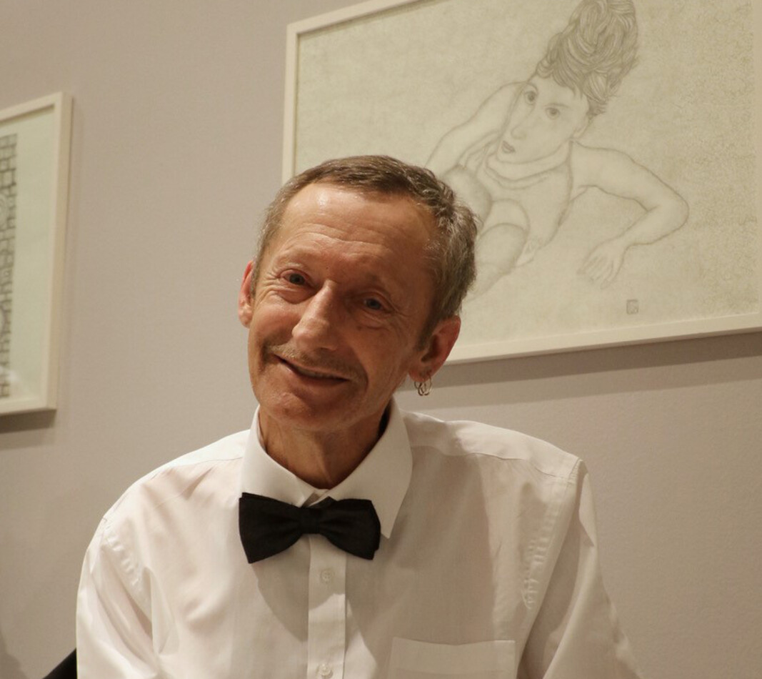 Ein Porträt des Künstlers Werner Roitner bei einer Ausstellung. Er sitzt lächelnd in seinem Rollstuhl und trägt ein elegantes weißes Hemd mit einer schwarzen Fliege. Im Hintergrund ist eines seiner Werke zu sehen: Eine gerahmte, feine Bleistiftzeichnung auf hellem Grund, die eine stilisierte menschliche Figur in einer dynamischen Pose zeigt. Die Szene vermittelt eine festliche und stolze Stimmung.