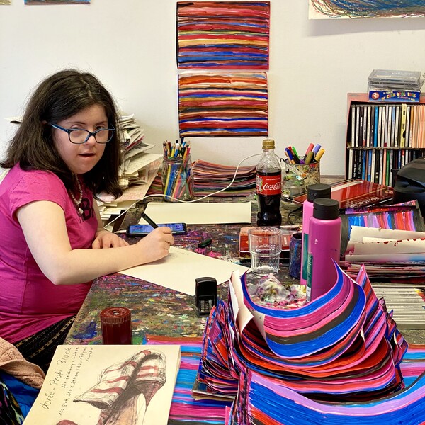 Andrea Meija Rocha Die Künstlerin Andrea Meija Rocha sitzt an ihrem Arbeitsplatz, sie hält einen Stift in der Hand, auf dem Tisch befinden sich verschiedene Kunstwerke, und Arbeitsmaterialien im Hintergrund an der Wand hängen Bilder