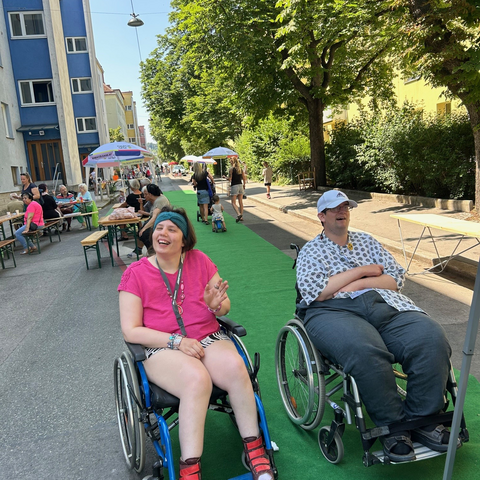 Grätzlfest Käthe-Dorsch-Gasse Zwei Bewohner im Rollstuhl lachen ausgelassen beim Grätzlfest in der Käthe-Dorsch-Gasse. Eine Frau im pinken T-Shirt und ein Mann im gemusterten Hemd mit weißer Kappe sitzen auf einem langen grünen Teppich, der mitten auf der Straße ausgerollt ist. Im Hintergrund der sonnigen, baumbestandenen Gasse sind Festbänke, Sonnenschirme und weitere Besucher zu sehen, die das Nachbarschaftsfest genießen.
