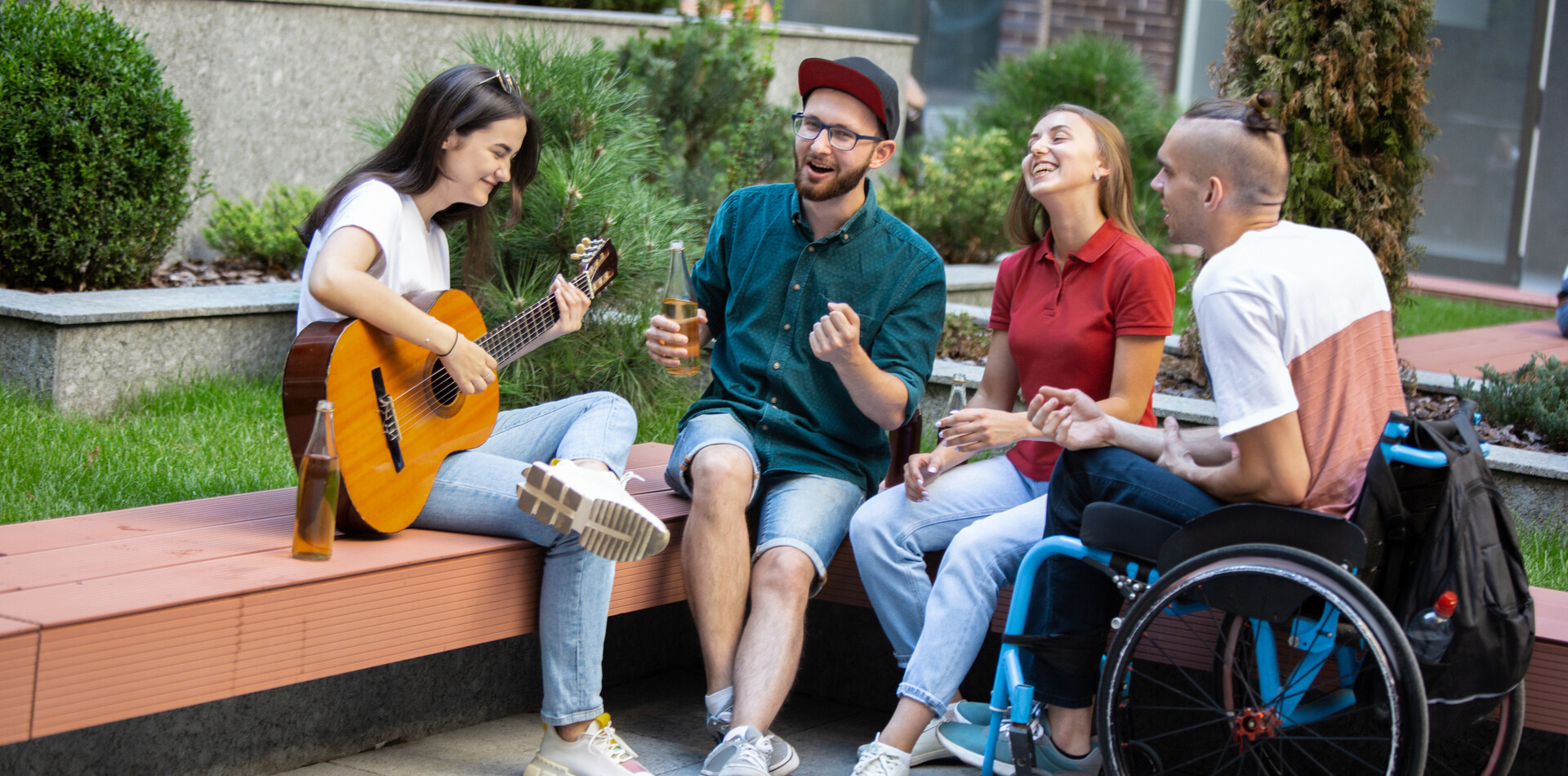 Teilbertreutes Wohnen im Wohnverbund Eine Gruppe junger Freunde, darunter ein junger Mann mit Rolstuhl sitzt lachend im Freien; eine Frau spielt Gitarre, während die anderen singen und Getränke halten.