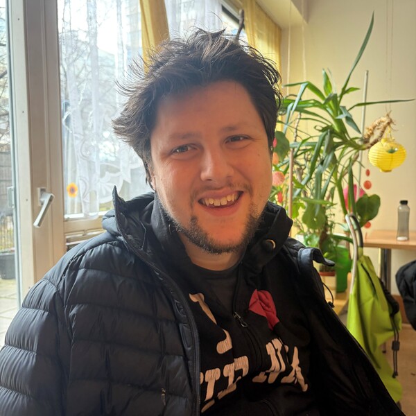 Bewohner Benedikt Ein lächelnder junger Mann mit dunklem Haar und schwarzer Steppjacke in einem hellen Raum mit Zimmerpflanzen.