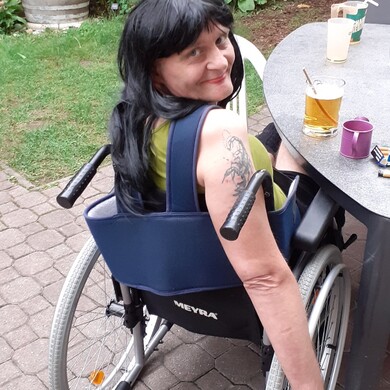 Eine Bewohnerin mit langen schwarzen Haaren und einem auffälligen Skorpion-Tattoo am Oberarm sitzt in ihrem Rollstuhl im Garten der Wohngemeinschaft Böckh. Sie trägt ein olivgrünes Top, hat die Lippen rot geschminkt und blickt mit einem verschmitzten Lächeln über die Schulter direkt in die Kamera. Sie befindet sich auf einer gepflasterten Terrasse an einem grauen Gartentisch, auf dem Gläser und Getränke stehen. Im Hintergrund ist eine grüne Gartenanlage mit Pflanzen und einem großen Holzfass zu sehen.