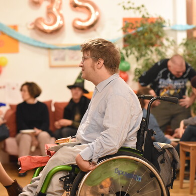 Auf einer Geburtstagsfeier in einer betreuten Wohngemeinschaft sitzen und stehen mehrere Personen mit und ohne Behinderung zusammen und unterhalten sich.