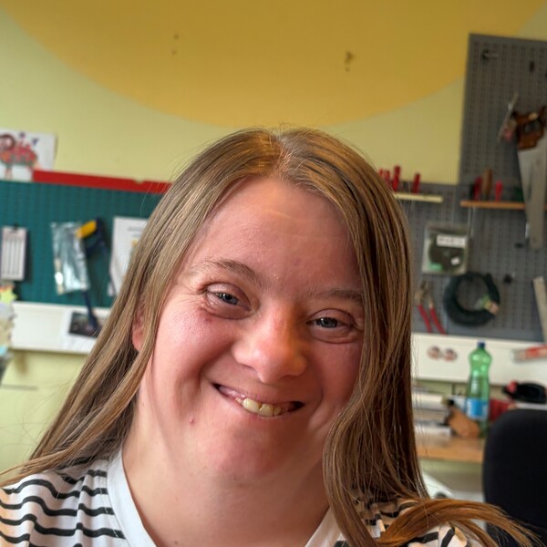 Tagesstruktur-SoHo-Testimonial-Suzana Porträtaufnahme von Suzana, einer Frau mit langen, braunen Haaren und einem herzlichen Lächeln. Sie trägt ein schwarz-weiß gestreiftes T-Shirt. Der Hintergrund zeigt eine helle Werkstatt mit Werkzeugwänden und einer gelben Decke.