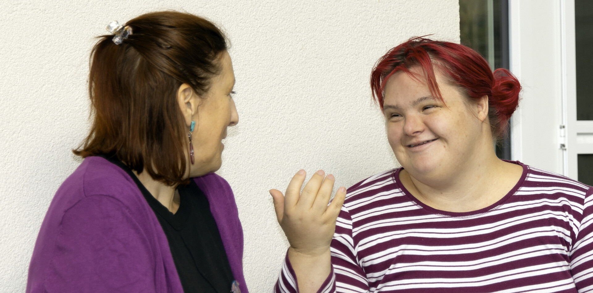 TAGS Käthe Dorsch I BALANCE Eine Frau mit braunen Haaren und lila Weste und eine Frau mit roten Haaren, die ein lila-weiß gestreiftes Oberteil trägt sind in einem Gespräch. Beide lächeln und wirken fröhlich und entspannt.