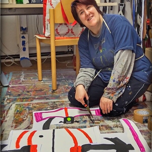 Die Künstlerin Barbara Plak kniet am Boden, hält eine Schere in der Hand und lacht in die Kamera, auf dem Boden vor ihr sieht man ein großes Kunstwerk aus Papier von dem ein Teil herausgeschnitten wurde.