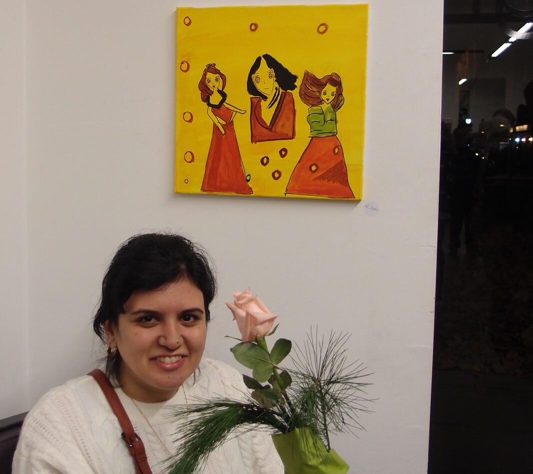 Juka Abdilwahid Die Künstlerin Juka Abdilwahid steht in einer Ausstellung vor einem ihrer Bilder und lächelt in die Kamera. rechts von ihr ist eine hellrosa Rose. Das Kunstwerk zeit drei Frauengestalten in roten Kleidern vor gelbem Hintergrund.