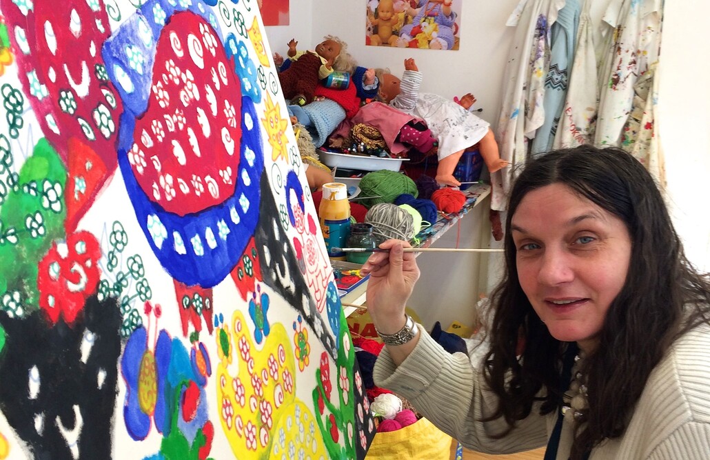 Eine Frau mit dunklen langen Haaren hält einen Pinsel in der Hand und malt an einem großen bunten Bild. Sie schaut in die Kamera.