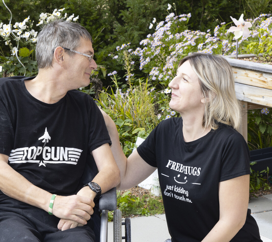 Im Außenbereich des Wohnverbunds Käthe Dorsch hockt eine BALANCE Mitarbeiterin neben einem Bewohner im Rollstuhl. Sie sehen sich an. Ein lächelnder Mann im Rollstuhl und eine Frau knien im Garten nebeneinander und schauen sich freundlich an. Der Mann trägt ein schwarzes T-Shirt mit dem „Top Gun“-Logo, die Frau ein schwarzes T-Shirt mit der Aufschrift „FREE HUGS – just kidding, don't touch me“. Im Hintergrund sind blühende Gartenpflanzen und ein Hochbeet zu sehen.