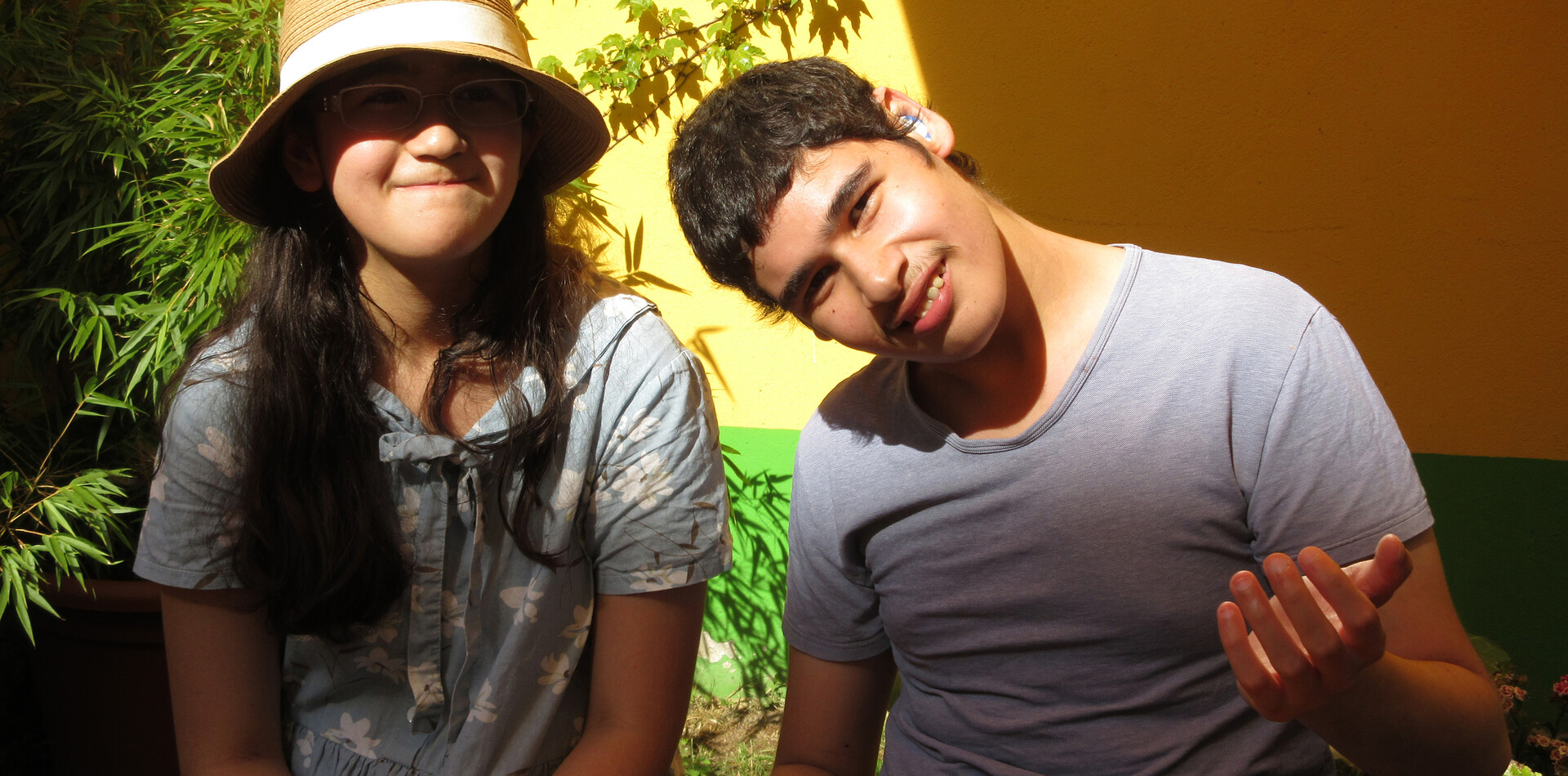 WG Salzach I BALANCE Eine junge Frau mit Sonnenhut und T-Shirt und ein junger Mann im T-Shirt sitzen im Hof der WG Salzach und lächeln in die Kamera