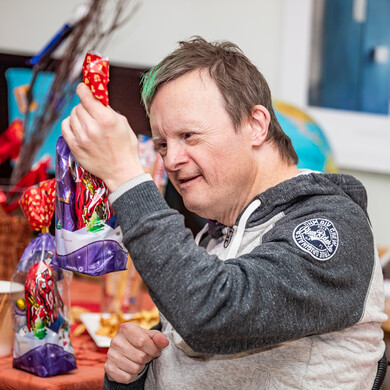 Wohnen bei BALANCE - Weihnachtsfeier Ein Mann mit Trisomie 21 und einer markanten grünen Haarsträhne blickt lächelnd auf eine lila-rote Geschenkverpackung, die er in der Hand hält. Er trägt einen sportlichen, grau-melierten Kapuzenpullover mit einem runden Logo-Aufnäher am Ärmel. Vor ihm auf einem Tisch stehen weitere weihnachtlich verpackte Geschenktüten. Der Hintergrund ist hell und leicht unscharf.