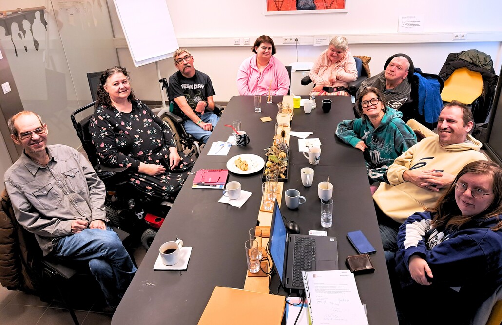 Das Wicky Team – die Ideen- und Beschwerdestelle für Nutzer:innen – bei einer gemeinsamen Besprechung an einem großen, dunklen Konferenztisch in der BALANCE Zentrale. Neun Personen sitzen in einer gemütlichen Runde zusammen, einige davon in Rollstühlen. Auf dem Tisch stehen Kaffeetassen, Gläser, ein Laptop und Unterlagen. Die Atmosphäre ist konzentriert und freundlich. An der weißen Wand im Hintergrund hängen ein Flipchart und ein farbenfrohes, abstraktes Kunstwerk.