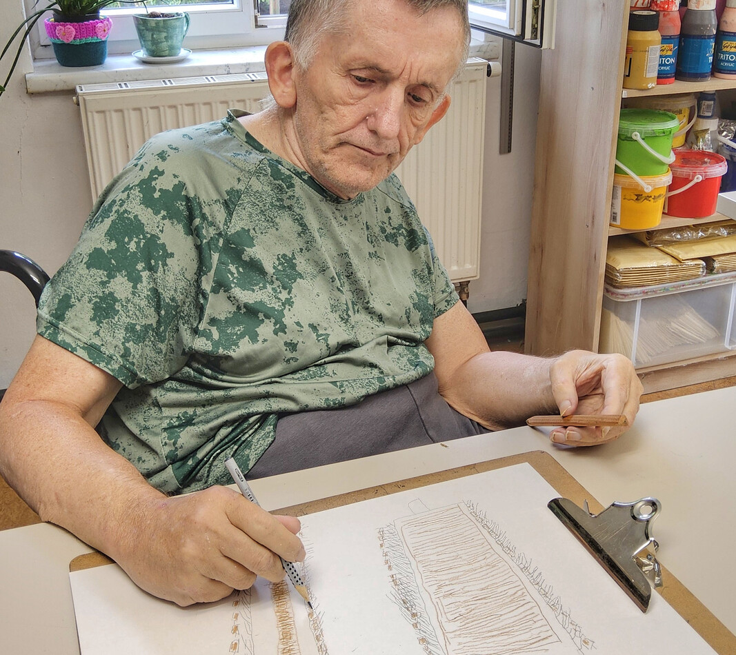 Ein Porträt des Künstlers Friedrich Ettenauer bei der Arbeit im Atelier Maria Ponsee. Er sitzt an einem Tisch vor einem geöffneten Fenster, durch das man ins Grüne blickt, und zeichnet konzentriert mit einem feinen Stift auf ein Klemmbrett. Vor ihm liegen zwei Skizzen, die feine, strukturierte Linien und handschriftliche Notizen zeigen. Rechts im Hintergrund steht ein Holzregal, das dicht mit bunten Farbtöpfen und Malutensilien gefüllt ist.