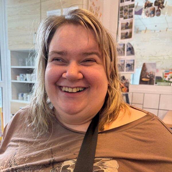 Tagesstruktur-Testimonial-Melanie Porträtfoto von Melanie, einer herzlich lachenden Frau mit hellem, schulterlangem Haar. Sie trägt ein braunes Oberteil mit floralen Mustern und einen schwarzen Umhängegurt. Der Hintergrund zeigt einen hellen, freundlichen Raum mit Regalen und Fotos an der Wand.