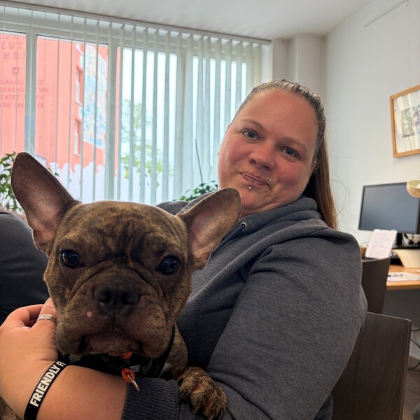Nutzerin Dani mit ihrem Hund Muffin Dani lächelt in die Kamera und hält Muffin, eine braun-schwarz gestromte Französische Bulldogge, auf dem Schoß. Sie befinden sich in einem hellen Büro. Dani trägt einen grauen Kapuzenpullover.