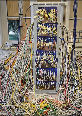 Eine raumfüllende Kunstinstallation von Barbara Plak im Innenbereich des Ateliers Maria Ponsee. Im Zentrum steht ein hoher, grauer Schaltschrank mit sechs blauen Fächern, aus denen hunderte bunte Kabel – vorwiegend in Weiß, Gelb, Blau und Rot – wie Tentakel hervorquellen. Die Kabel ergießen sich in einem gewaltigen Wirrwarr über den Boden und füllen den unteren Bereich des Bildes fast vollständig aus. Einige Kabelstränge führen geordnet an den Seiten nach oben. Im Hintergrund sind ein Fenster und ein Teil einer Wandzeichnung mit einer Roboterhand zu sehen.