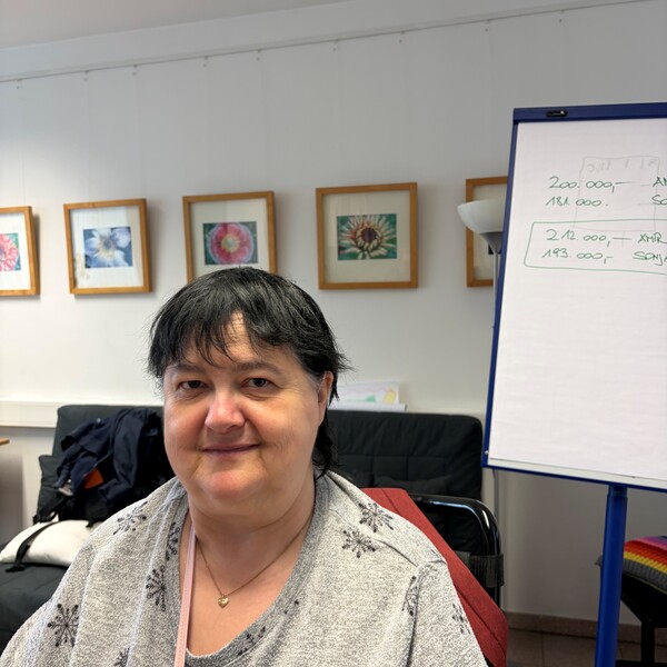 testimonial-mobi12-helga Porträt einer leicht lächelnden Frau mit kurzen, dunklen Haaren (Helga), die ein graues Oberteil mit schwarzen Schneeflocken-Mustern und eine feine Halskette trägt. Sie sitzt in einem hellen Raum; im Hintergrund hängen gerahmte Bilder an der Wand und rechts steht ein Flipchart mit handschriftlichen Notizen.