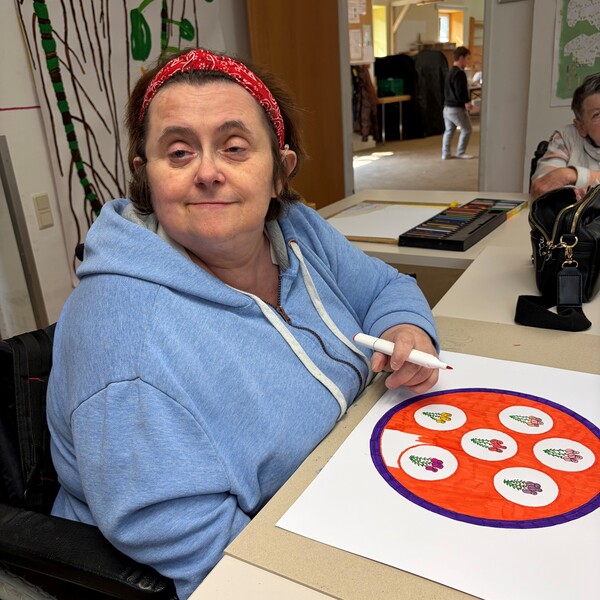 Die Künstlerin Gabi Mühlbauer mit einem roten Haarband und einem hellblauen Kapuzenpullover sitzt lächelnd an einem Tisch und zeichnet. Vor ihr liegt ein weißes Blatt Papier, auf dem ein rundes, farbenfrohes Kunstwerk mit Blumenmotiven in Rot- und Lilatönen zu sehen ist. Sie hält einen weißen Filzstift in der Hand; im Hintergrund sind ein Malkasten und weitere Zeichnungen erkennbar.