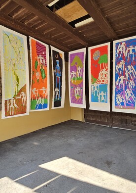 Eine Kunstinstallation von Barbara Plak im überdachten Außenbereich des Ateliers Maria Ponsee. Acht meterhohe, vertikale Papierbahnen hängen nebeneinander an einer Holzbalkendecke und bilden eine bunte Galerie. Jede Bahn zeigt eine fantasievolle Szene mit den für die Künstlerin typischen weißen, roboterartigen Figuren (Aqadroiden). Die Hintergründe variieren in kräftigen Farben wie Blau, Orange, Pink, Grün und Gelb und stellen unterschiedliche Landschaften dar – von Unterwasserwelten mit Algen bis hin zu Szenen unter roten Planeten.