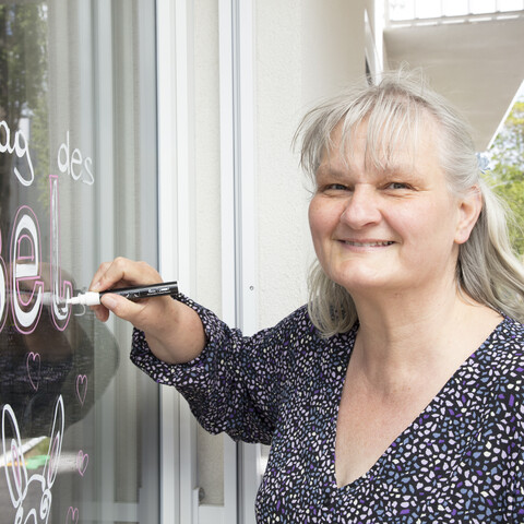 Tagesstruktur Käthe Dorsch - Monatsaffirmation Eine Teilnehmerin der Tagesstruktur Käthe Dorsch steht lächelnd vor einer Glasscheibe und schreibt mit einem weißen Kreidemarker die Worte ‚Welttag des Esels‘ auf das Fenster. Unter dem Schriftzug sind eine kleine Esel-Illustration und Herzen zu sehen. Die Frau mit grauem Haar trägt eine dunkle Bluse mit feinem Muster und blickt freundlich in die Kamera.