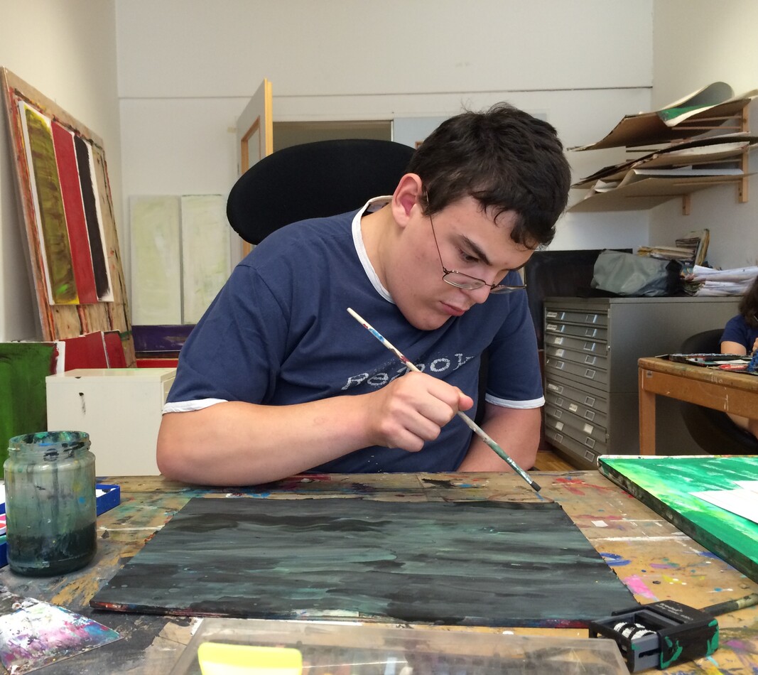 Darko Beric Der Künstler Darko Beric sitzt an seinem Arbeitspaltz und malt an einem Bild in grün und schwarzen Farbtönen. Er blickt konzentriert auf das Blatt vor ihm. Er dunkle kurze Haare und trägt ein blaues T-Shirt. Im Hintergrund sieht man fertige Kunstwerke an der Wand lehnen.