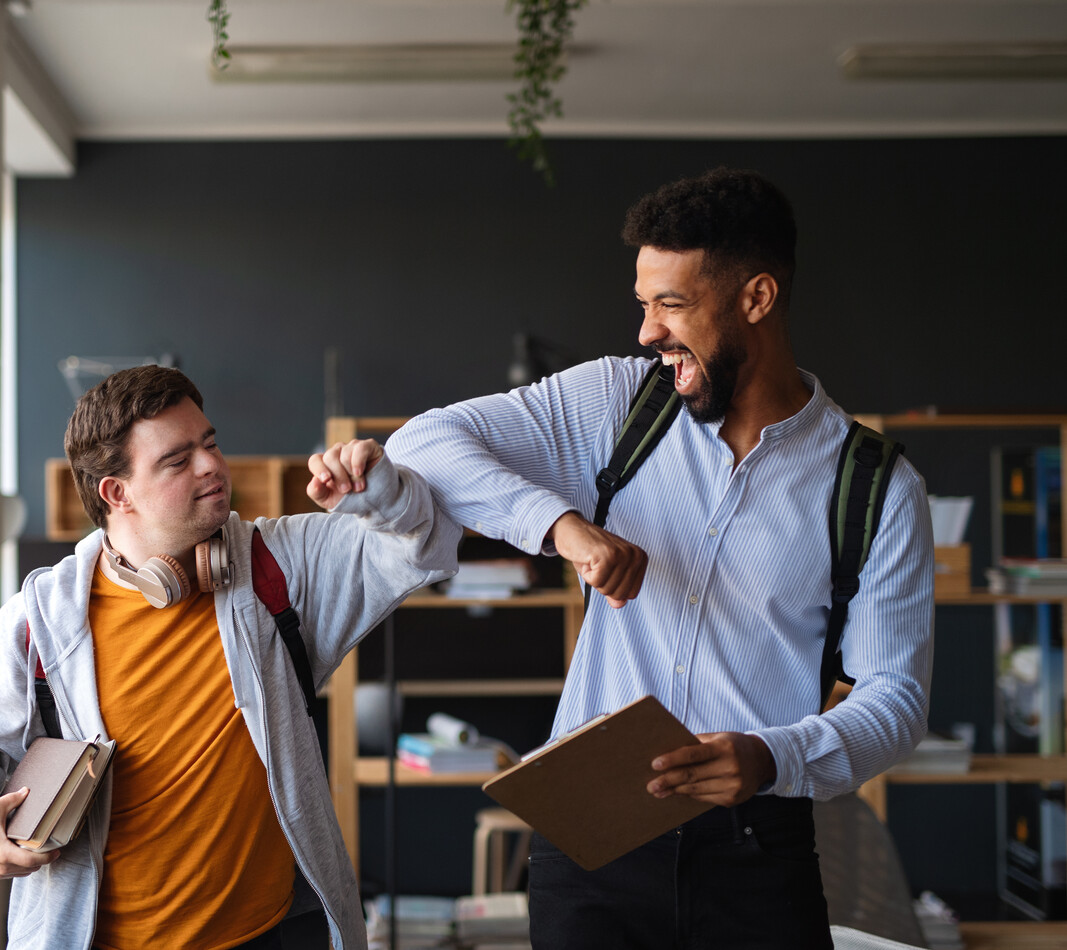 Jobs bei BALANCE Zwei junge Männer begrüßen sich in einem hellen, modernen Innenraum mit einem freundschaftlichen Ellenbogencheck. Beide tragen Rucksäcke und halten Bücher bzw. ein Klemmbrett in den Händen. Der linke Mann mit Trisomie 21 lächelt leicht, während der rechte Mann mit dunkleren Locken und Bart herzlich lacht. Die Szene vermittelt eine Atmosphäre von Freude, Gemeinschaft und gelebter Inklusion.