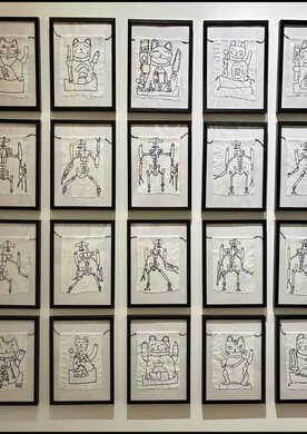 Wandinstallation der Künstlerin Barbara Plak aus der 20-teiligen Bild-Serie „Gebetsfahnen „. In einem Raster von vier Reihen und fünf Spalten hängen 20 schwarz gerahmte Bilder an einer weißen Wand. Jedes Bild zeigt eine feine, schwarze Linienzeichnung auf weißem Stoff. Die oberste und unterste Reihe zeigen Variationen der winkenden Glückskatze (Maneki-neko), während die beiden mittleren Reihen verschiedene Roboter-Figuren (Aquadroiden) in dynamischen Posen darstellen.
