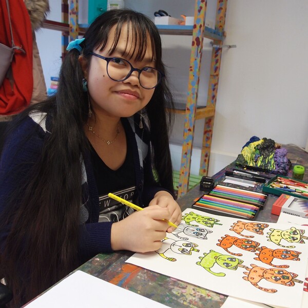 Ashley Horney Die Künstlerin Ashley Horney sitzt an ihrem Arbeitsplatz und zeichnet auf ein Blatt Papier. Sie lächelt in die Kamera. Sie hat lange dunkle Haare, trägt eine Brille und ein dunkelblaues Shirt mit weißem Schriftaufdruck.