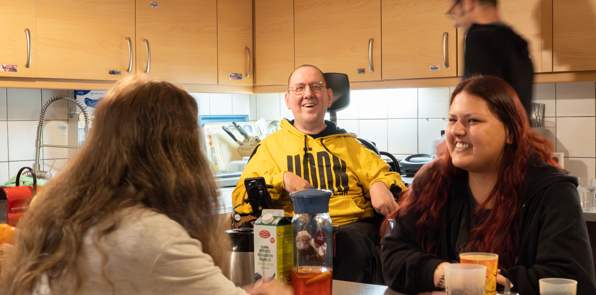 Gemeinschaftsküche der WG Böckh In der Wohngemeinschaft Böckh sitzen drei Personen gemeinsam an einem Tisch in einer hellen Küche und unterhalten sich gut gelaunt. Ein Mann im gelben Kapuzenpullover, der einen Rollstuhl nutzt, blickt strahlend in die Kamera. Ihm gegenüber sitzt eine junge Frau mit langen rötlichen Haaren, die ihn lächelnd ansieht. Im Vordergrund ist eine weitere Person von hinten zu sehen. Auf dem Tisch stehen Getränke und eine Packung Milch. Im Hintergrund sind helle Küchenschränke und eine weitere Person in Bewegung erkennbar.