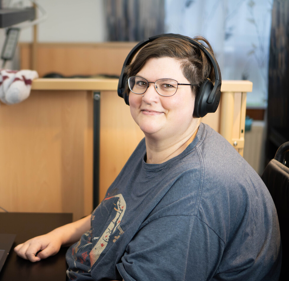 Person mit Kopfhörern sitzt an einem Schreibtisch und lächelt in die Kamera – eine Alltagssituation beim selbstständigen Wohnen.