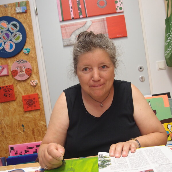 Bettina Onderka Die Künstlerin Bettina Onderka sitzt an ihrem Arbeitsplatz im bildBalance Atelier Wien und blickt mit einem sanften Lächeln in die Kamera. Sie trägt ein schwarzes, ärmelloses Oberteil und hat ihr graumeliertes Haar hochgesteckt. Vor ihr auf dem Tisch liegt eine aufgeschlagene Zeitschrift und ein grünes Bild, an dem sie gerade arbeitet. Die Wand hinter ihr ist mit vielen ihrer farbenfrohen, quadratischen und runden Kunstwerke auf Holz und Leinwand geschmückt. Rechts im Bild ist ein Regal mit Malutensilien und einer grünen Stofftasche zu sehen.