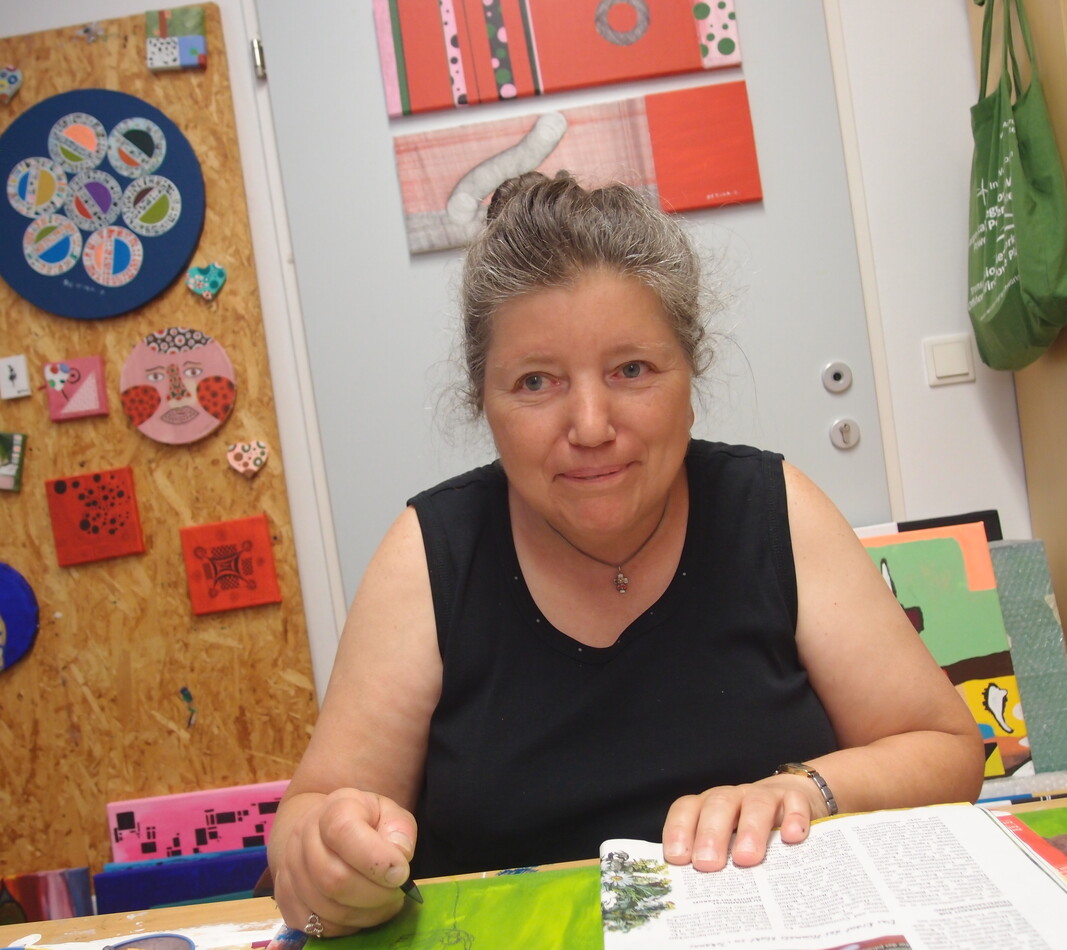 Bettina Onderka Die Künstlerin Bettina Onderka sitzt an ihrem Arbeitsplatz, sie trägt ein ärmelloses schwarzes Kleid, ihre Haare sind hochgesteckt. Sie schaut gerade in die Kamera, im Hintergrund sieht man verschiedene Kunstwerke.