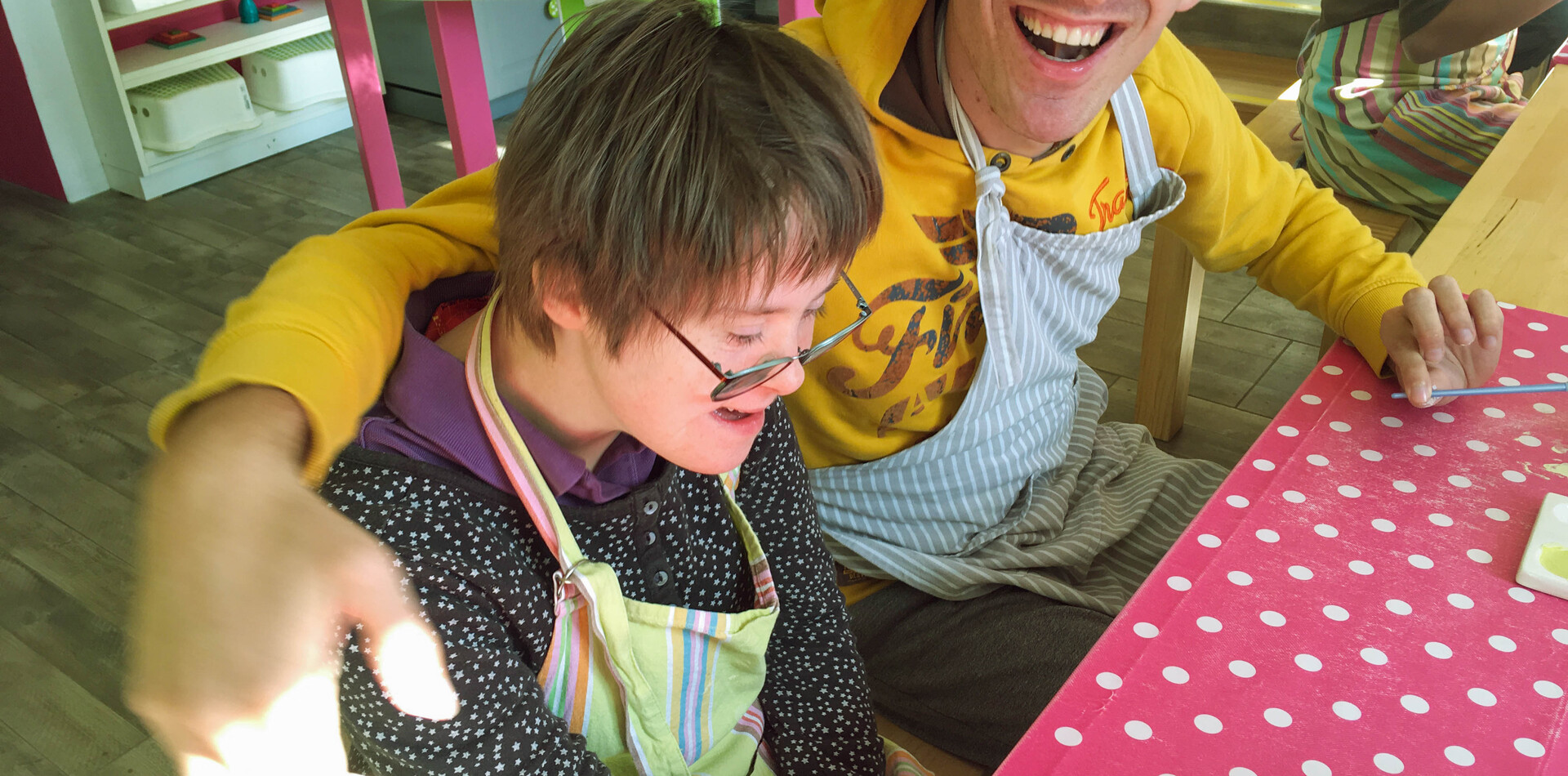 TAGS SoHo I BALANCE Ein junger Mann und eine junge Frau sitzen an einem Tisch mit einer roten Tischdecke mit weissen Punkten. Sie tragen beide Arbeitsschürzen und lachen. Der Mann hat seinne Arm um die Schultern der Frau gelegt. Beide lachen.