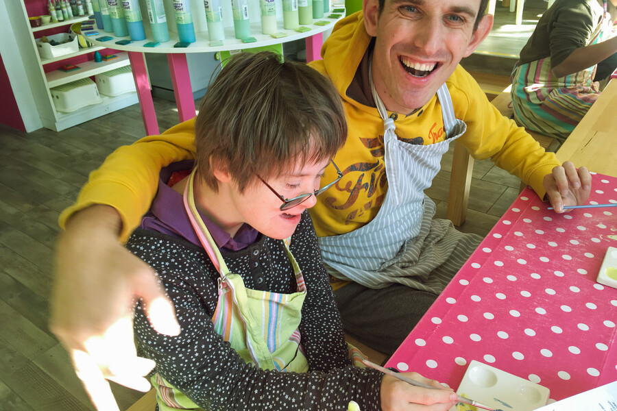TAGS SoHo I BALANCE Ein junger Mann und eine junge Frau sitzen an einem Tisch mit einer roten Tischdecke mit weissen Punkten. Sie tragen beide Arbeitsschürzen und lachen. Der Mann hat seinne Arm um die Schultern der Frau gelegt. Beide lachen.