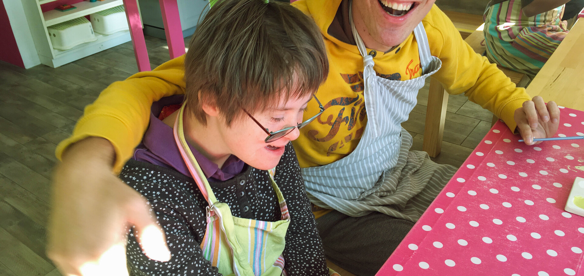 TAGS SoHo I BALANCE Ein junger Mann und eine junge Frau sitzen an einem Tisch mit einer roten Tischdecke mit weissen Punkten. Sie tragen beide Arbeitsschürzen und lachen. Der Mann hat seinne Arm um die Schultern der Frau gelegt. Beide lachen.