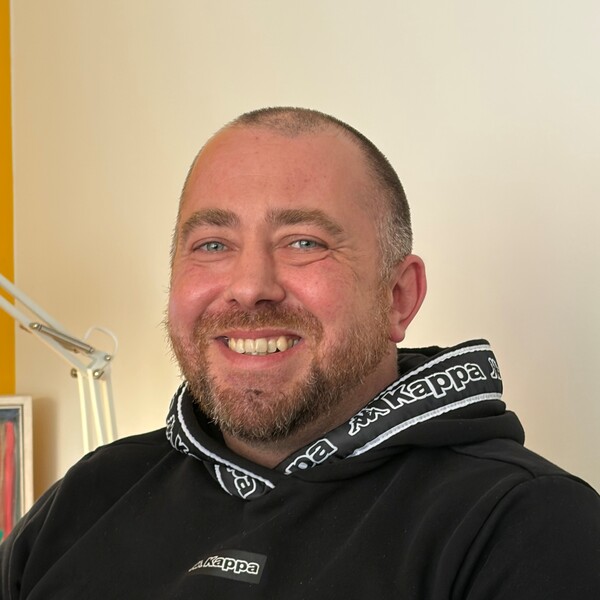 BALANCE Mitarbeiter Andi aus dem Team der Mobi Zwölf Mitarbeiter Andi sitzt entspannt in einem Innenraum. Er trägt einen schwarzen Hoodie mit weißen Kappa‑Logos am Kragen und auf der Brust. Im Hintergrund sind eine gelbe Wand, eine Lampe und ein gerahmtes Bild zu sehen.