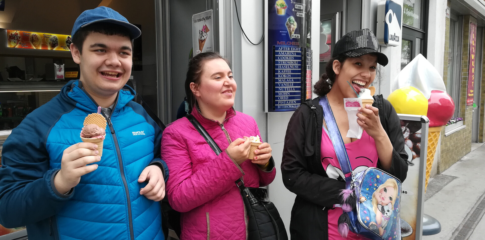 TAGS Auhof I BALANCE Ein jungern Mann und zwei Frauen stehen vor einem Eisgeschäft. Sie haben alle jeweils ein Eis in der Hand und lachen.