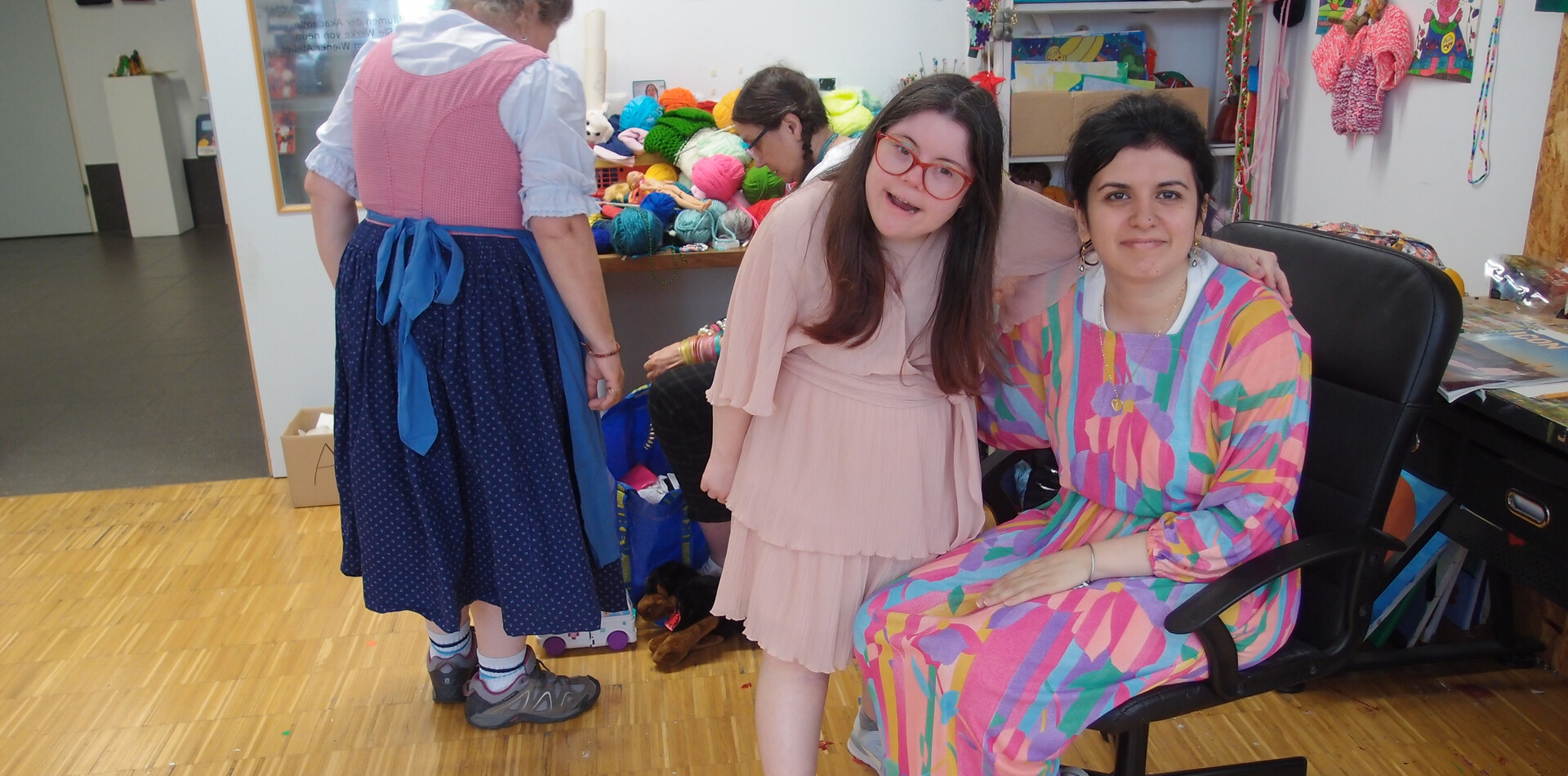 TAGS Fuchsenfeld I BALANCE Zwei lächelnde Personen in einem kreativen Arbeitsraum: im Vordergrund sitzen eine Person in einem rosafarbenen Kleid und eine Person in einem farbenfrohen, gemusterten langen Kleid sitzt auf einem Bürostuhl. Beide lächeln in die Kamera und haben den Arm um ihre Schultern gelegtIm Hintergrund sind Handarbeitsmaterialien wie Wolle und Bastelutensilien zu sehen, eine Person im Dirndl, sie steht mit dem Rücken zur Kamera und spricht mit einer anderen sitzenden Person,