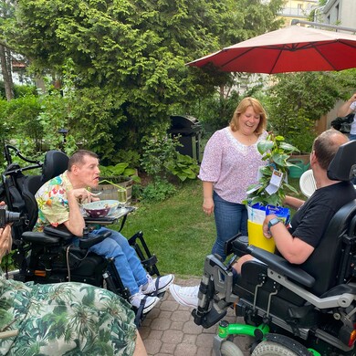 Mehrere Menschen mit und ohne Behinderung sitzen und stehen in einem Garten zusammen. Eine Frau bekommt eine Topfpflanze als Geschenk überreicht.