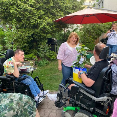 Mehrere Menschen mit und ohne Behinderung sitzen und stehen in einem Garten zusammen. Eine Frau bekommt eine Topfpflanze als Geschenk überreicht.