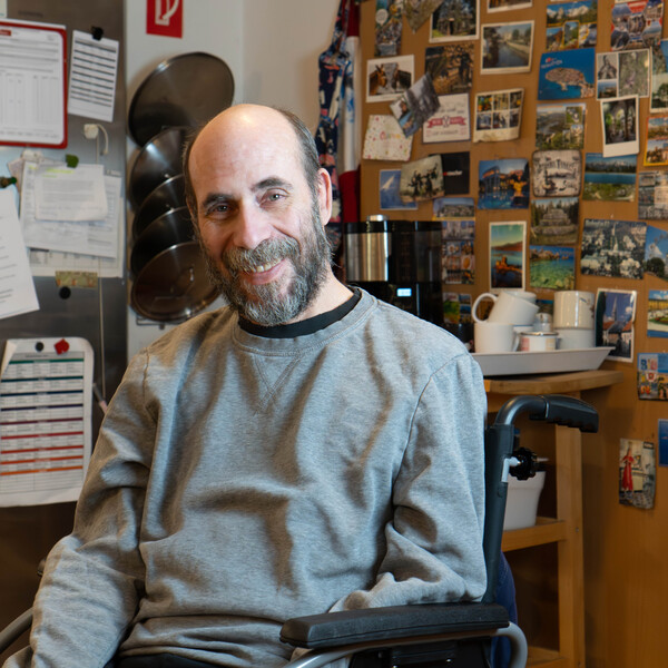 Bewohner und Rollstuhlfahrer Harald lächelt in die Kamera, hinter ihm eine Wand mit vielen Ansichtskarten.