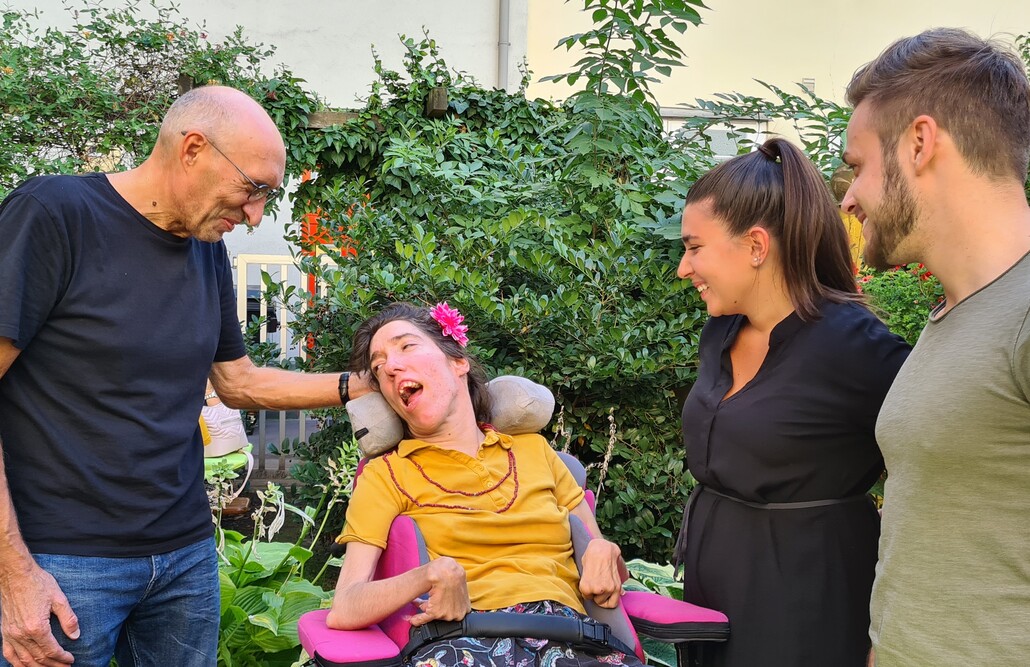 Bei einem Gartenfest stehen und sitzen vier Personen mit und ohne Behinderung nebeneinander und unterhalten sich.